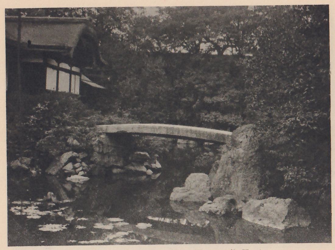 Photograph of a pond with floating leaves in the foreground, a low arched stone bridge across it at the center, large rocks at the water’s edge on the right and left, and a thatched building with pale paper windows at the upper left among dense trees.
