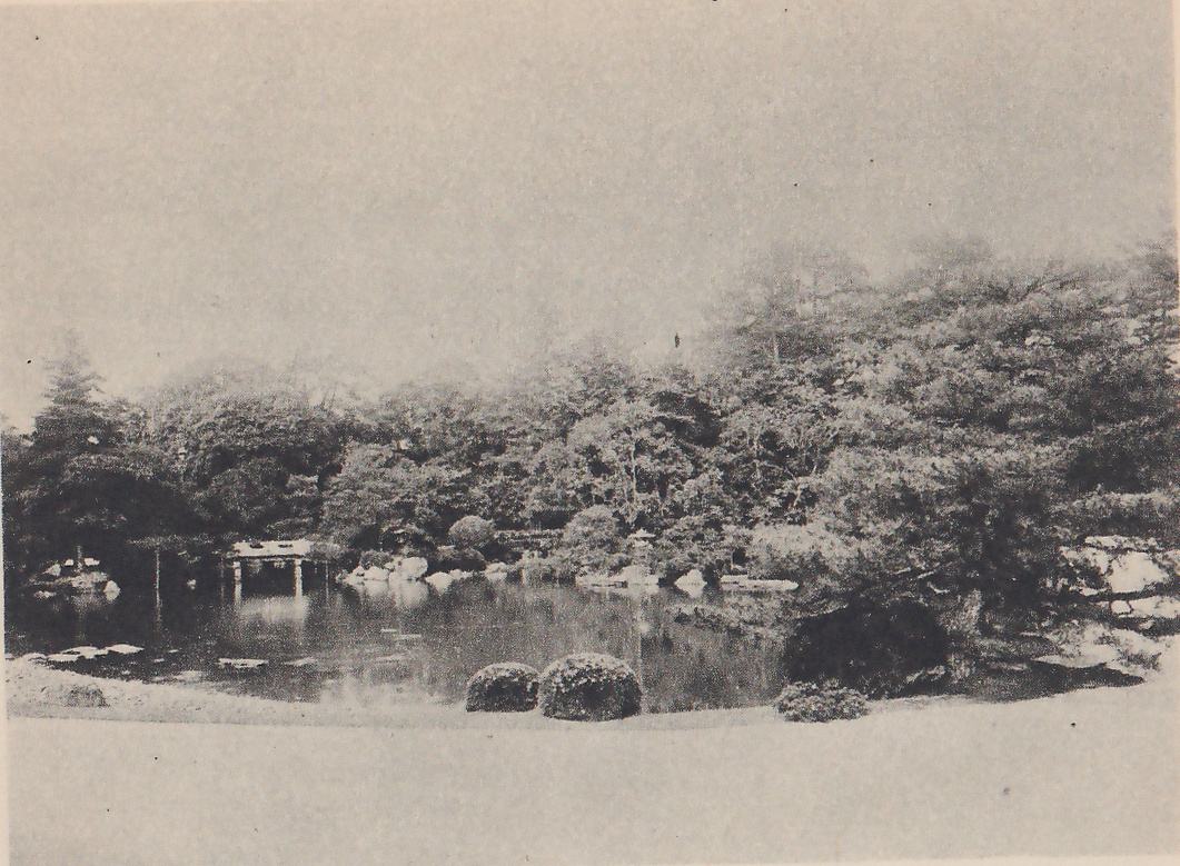 Photograph of a pond lying behind a wide bare band of pale gravel in the foreground, with round clipped shrubs at its near edge, a small roofed bridge or pavilion on the far shore at the left, and pines and other trees massed behind it under a pale sky.