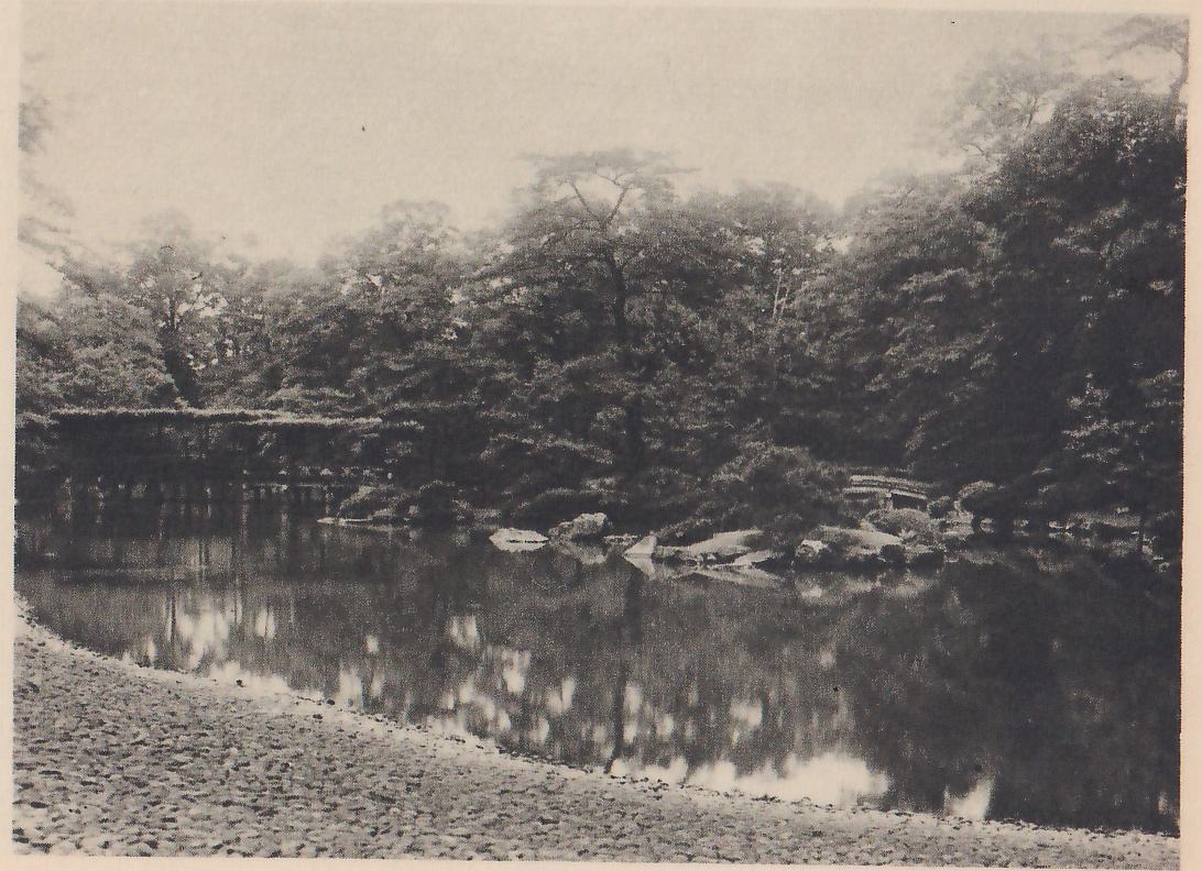 Photograph of a dark pond bordered in the foreground by a wide curving beach of small pebbles, with a long low trellised structure on posts over the water at the left, rocks and a small arched bridge on the far bank at the right, and dense trees behind.