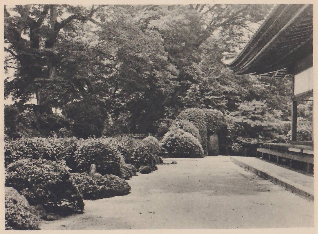 Photograph of a wide band of bare pale gravel running back between rounded clipped shrubs at the left and a raised wooden verandah under a large, deep tiled roof at the right, with tall trees behind and a small upright stone among the shrubs at the center.