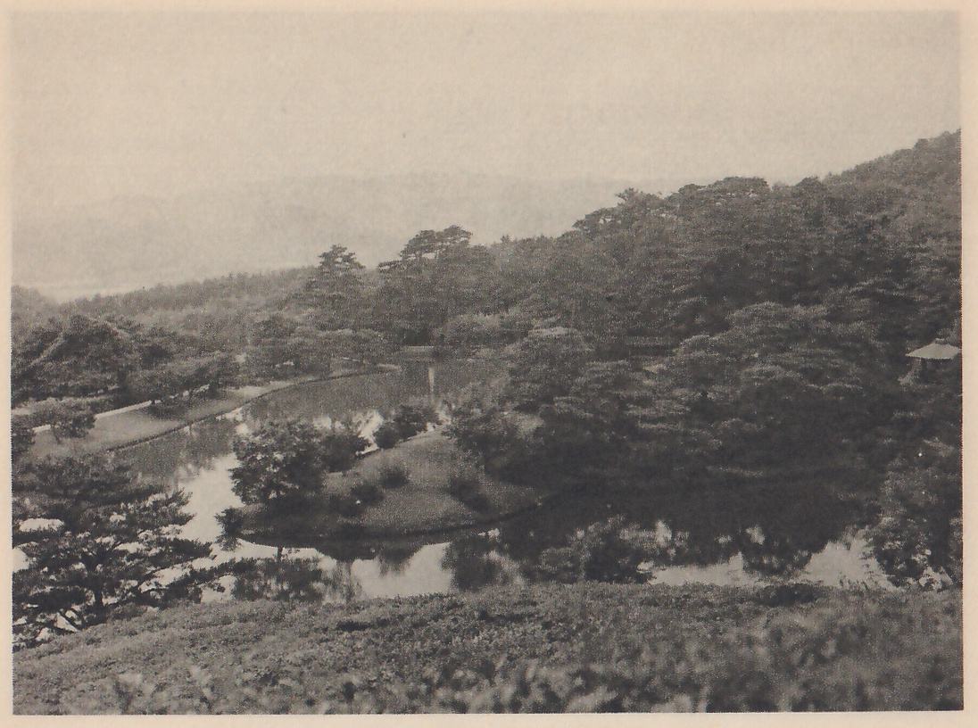 Photograph from a height over a garden: a grassy slope of low clipped shrubs in the foreground, a pond with a tree-covered island and a long narrow inlet beyond it, a mass of pines on a hillside at the right with a small roofed object at its edge, and hazy hills at the horizon.