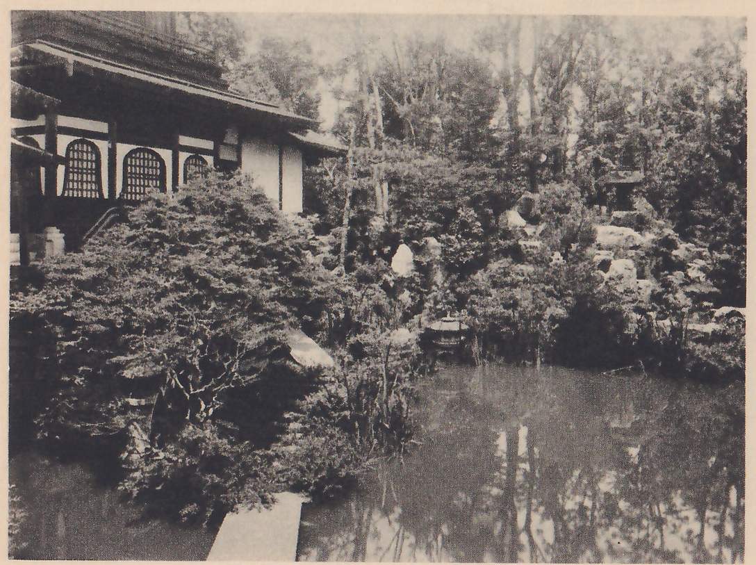 Photograph of a still pond in the foreground with a flat stone slab at its lower left edge, a large clipped evergreen at the left in front of a building with arched lattice windows and a deep eave, and rocks, a stone lantern and trees on the far bank.