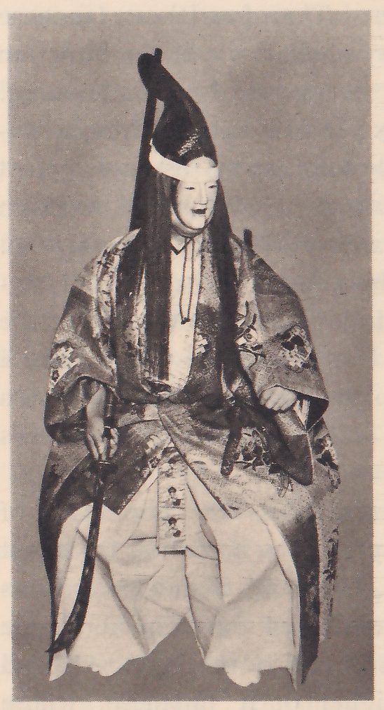 Photograph against a plain gray ground of a seated costumed figure in a pale mask facing the viewer, with a tall black pointed cap with a white band, long black hair hanging down on both sides of the face, a dark patterned outer robe over a white garment and wide white trousers; the hand at the viewer's left holds a long curved sword that reaches nearly to the floor, and the other hand rests on the lap.