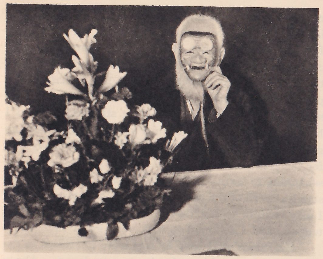 Photograph of a man in dark clothing, named by the printed caption as Mr. Bernard Shaw, seated at the right behind a pale cloth-covered surface; a mask with a white fringe of hair and beard covers his face and one hand is raised to its chin. At the left in the foreground a shallow dish holds a tall arrangement of lilies and other flowers.