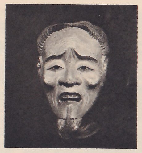 Photograph of a pale mask of an old face on a dark ground: a wrinkled forehead and cheeks, drooping lids, an open mouth, wispy hair at the temples and a thin beard hanging below the chin.