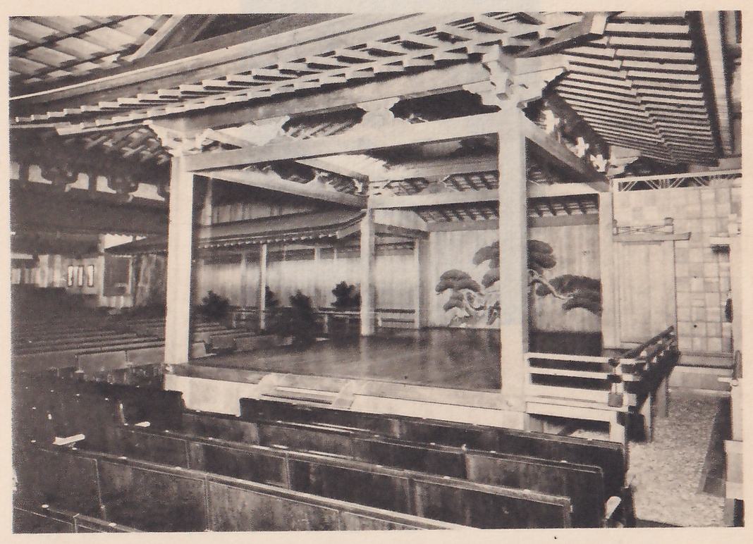 Photograph, taken from among the rows of seats, of a roofed wooden stage on thick pillars with a pine tree painted on its back wall; a railed walkway runs in along the right side, a strip of pebbles lies at the lower right, and rows of seats fill the foreground and the left.