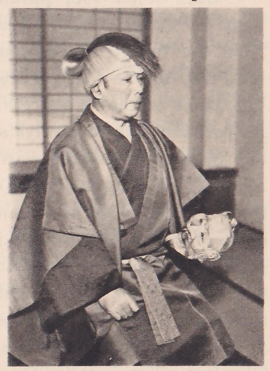 Photograph of a man seated in profile, facing the viewer's right, in a dark wide-shouldered robe, with a band around his head and a dark cloth on top; a pale mask shows beside his lap, and a paper-paned screen fills the upper left.