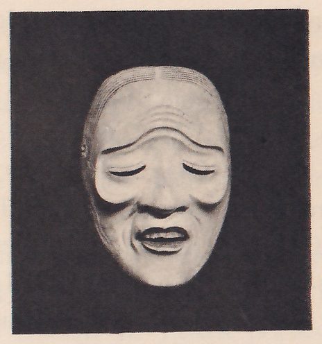 Photograph of a pale mask on a dark ground: a forehead ridged with curved lines, lowered eyelids, lined cheeks and a small, slightly open mouth.