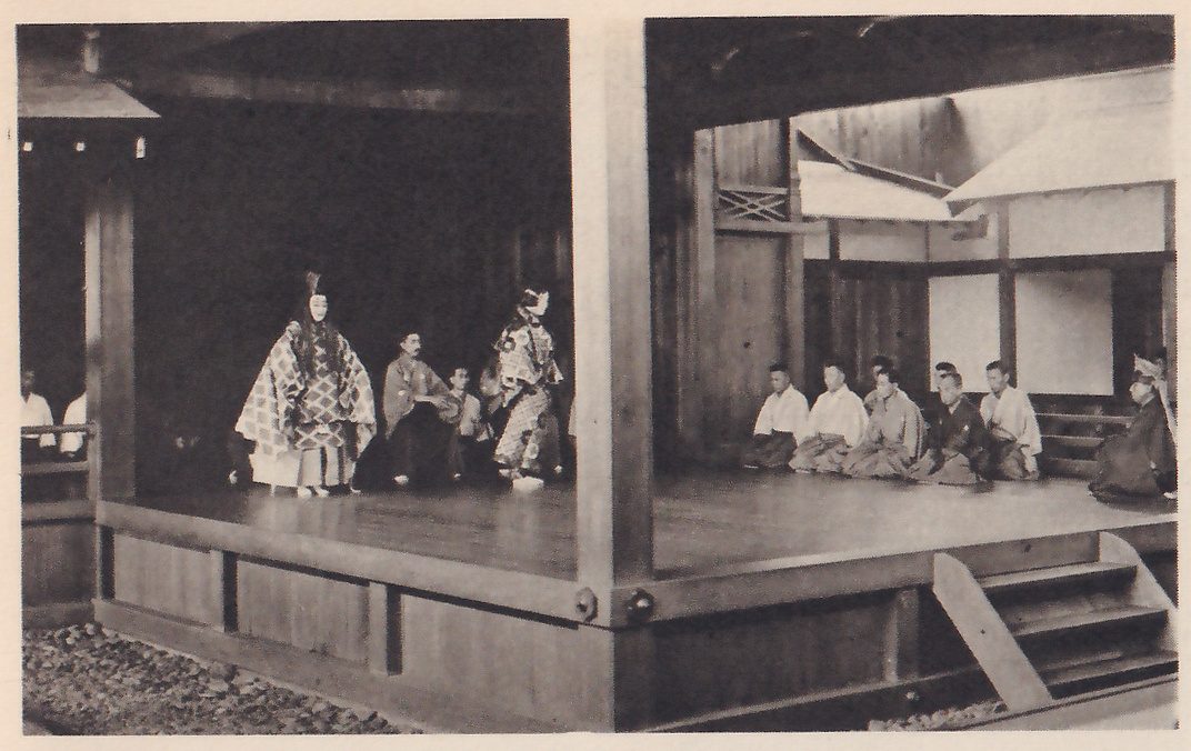 Photograph of a wooden stage under a roof, divided by a thick pillar: at the left a standing figure in a mask and a robe with a diamond pattern, a seated man, and a second standing figure in a patterned robe; at the right a row of men in pale and dark robes sit side by side along the edge of the stage. Wooden steps lead down at the lower right and pebbles cover the ground at the lower left.