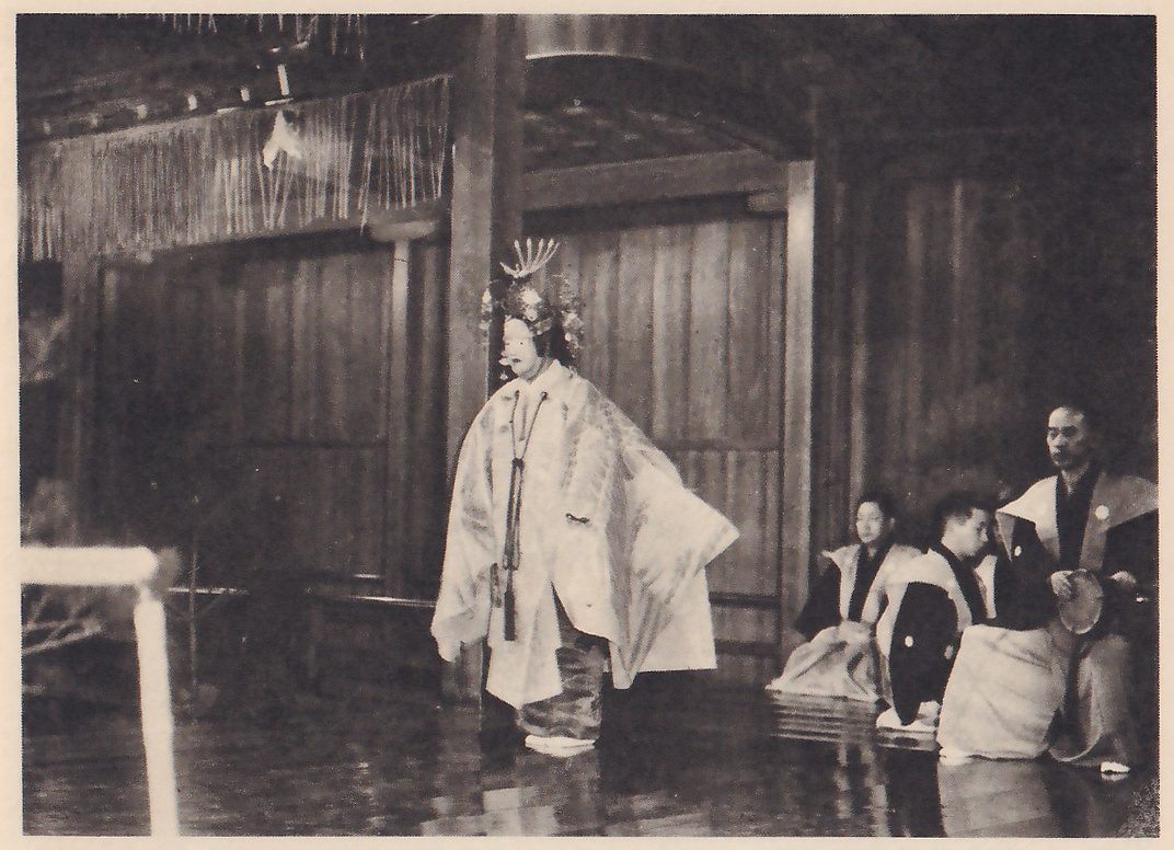 Photograph of a stage: at the left of center a masked figure in a tall ornate crown with a spiky ornament on top stands in a long pale robe with wide sleeves and a cord hanging down the front; at the right several people in dark and pale clothes sit on the floor, the one at the far right holding a round object at the hip. Wooden panel walls rise behind them, with a fringed roof edge at the upper left and a low railing at the lower left.