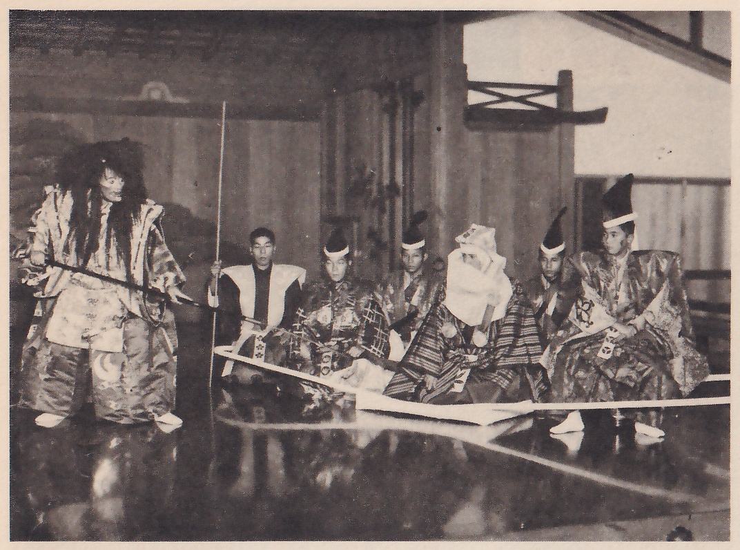Photograph of a stage: at the left a standing figure with very long loose dark hair and a pale mask, in a patterned robe, holds a long dark pole across the front of the body; in the middle and at the right sit men in patterned robes, several wearing tall pointed black caps with white bands and one in a white head covering bent forward, and a man in the middle holds a tall thin pole upright. Long pale strips of a light frame lie across the floor in front of them.