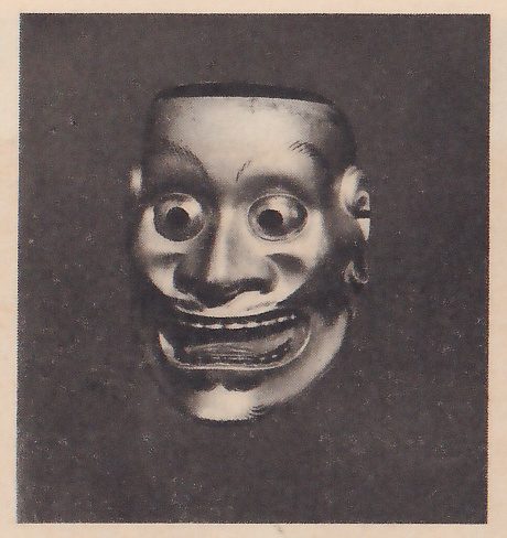Photograph of a dark mask on a dark ground: bulging round eyes with dark pupils, a broad nose, a wide mouth open in a grin that shows a row of teeth, lines across the cheeks and a rough chin.