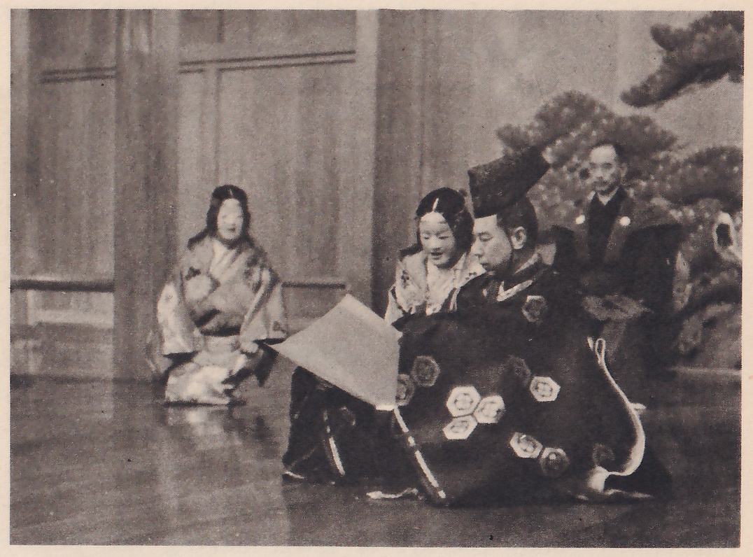 Photograph of a stage in front of a painted pine tree: at the right a man in a tall black cap and a dark robe with a pattern of white hexagons sits holding a large sheet of paper out toward the viewer's left; two figures in masks and pale patterned robes are with him, one kneeling at the left of the picture, and a man in dark clothes appears behind him at the right.