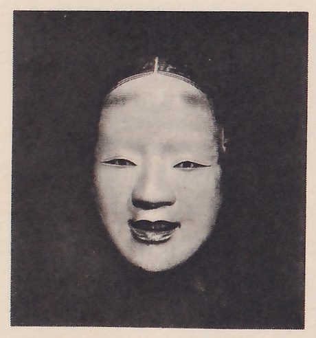 Photograph of a pale mask on a dark ground: a smooth rounded forehead with a thin line at the top where the hair parts, narrow eyes, a small nose and a small mouth, slightly open.