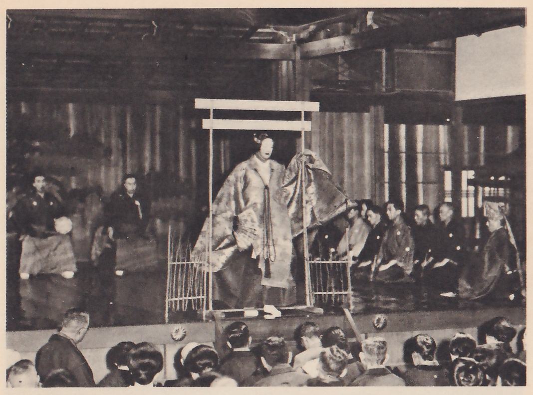 Photograph, taken from among the audience, of a stage: a masked figure in a long pale patterned robe with a cord hanging down the front stands inside a light frame with two crossbars on top and low fence panels at its sides; at the right a row of men in dark robes sit on the floor, and at the left two men in dark robes kneel. The heads of many spectators fill the bottom of the picture.