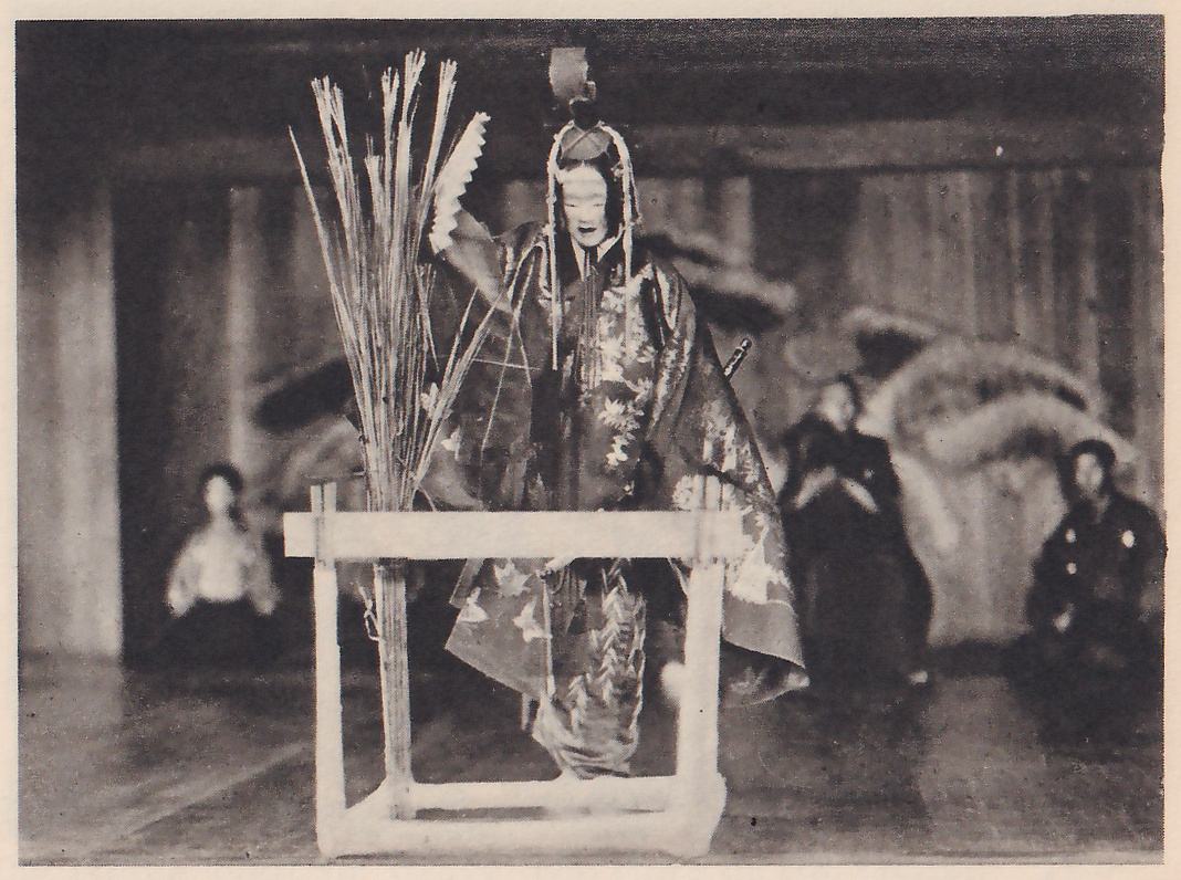 Photograph of a stage: at the center a masked figure in a headdress with hanging strands and a dark robe patterned with pale leaves stands behind a low white wooden frame, with a bundle of tall dry grass stalks rising at the left of the frame; one arm is raised toward the viewer's left with a pale fan beside the hand. Two seated people in dark clothes sit at the right and one in a pale top at the left.