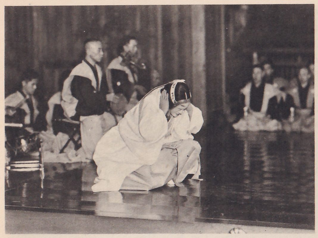 Photograph of a stage: in the foreground a person in a pale head covering and a loose pale robe kneels low on the polished floor, head bowed, with hands held up beside the head; behind at the left a man in a dark jacket with a pale shoulder cloth stands near two seated men, and at the upper right several men in dark clothes sit in a row. The floor reflects the figures.
