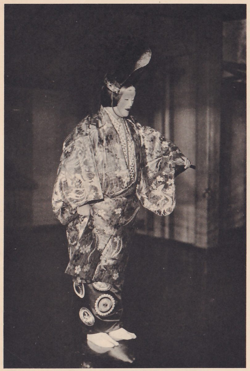 Photograph of a standing masked figure turned toward the viewer's right against a dark room: a tall dark headdress with a pale curved point rising behind the head, wide-sleeved robes covered with a pattern of flowers and birds, one arm held out toward the viewer's right with the hand around a thin dark rod, a dark lower garment with large white circles near the hem, and white split-toe socks.