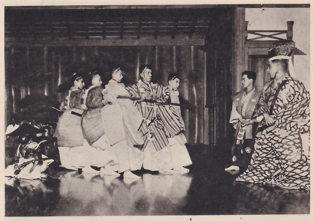 Photograph of a stage: at the left a row of figures in pale and striped robes lean toward the right, holding a long dark pole level between them; at the right, facing them, a figure in a patterned robe and a tall headdress with a white band kneels with a hand on a sword, and another figure stands beside it. A small figure crouches on the floor at the far left.