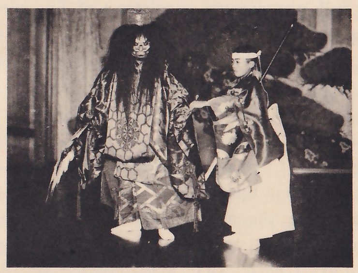 Photograph of two costumed figures on a stage in front of a painted pine tree: at the left a taller figure with a mask and very long loose dark hair, in robes with large circular patterns; at the right a smaller figure with a white headband and a patterned robe reaches an arm toward it, with a thin pole rising behind the shoulder.
