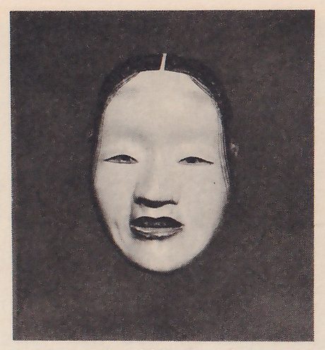 Photograph of a pale mask on a dark ground: dark hair parted down the middle with a light line at the top, narrow eyes, a small nose and dark lips, slightly open.