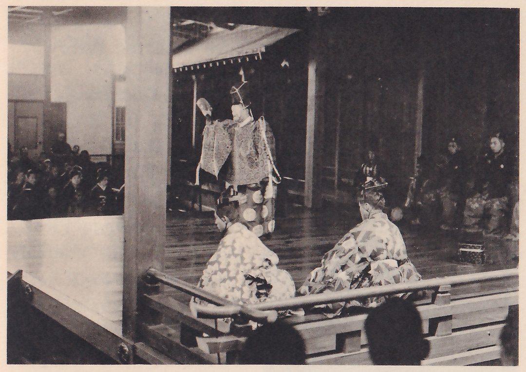 Photograph taken beside a wooden railing at the edge of a stage: a masked figure in a tall pointed hat and a stiff pale robe with a round pattern stands at the back left with one arm raised, and in the foreground two seated figures in robes with a pale angular pattern sit with their backs partly to the viewer. A thick wooden pillar stands at the left, with a crowd of spectators beyond it behind a white partition, and several men in dark clothes sit at the right of the stage.