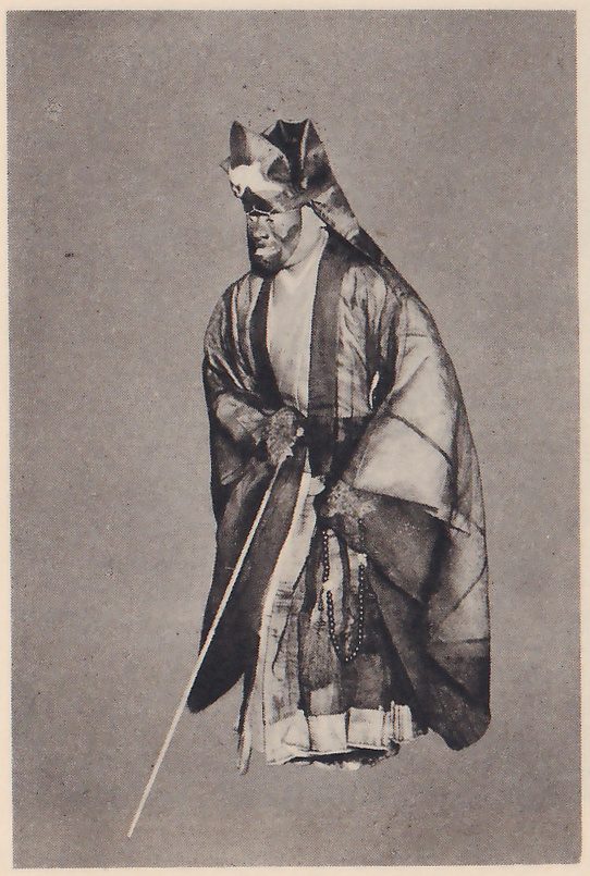 Photograph against a plain gray ground of a standing figure in a long, loose dark robe over a pale garment, with a cloth wrapped over the head into points on top, a dark face with a short beard, a long thin staff slanting down in the hand at the viewer's left, and a string of beads hanging at the waist.
