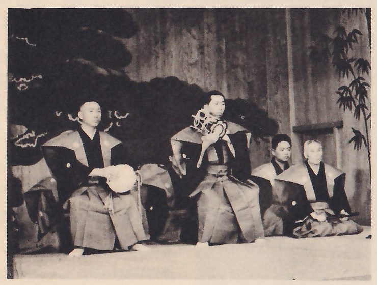 Photograph of men in dark robes with wide shoulder cloths sitting in a row on a low stage in front of a painted pine on a wooden wall, with bamboo painted at the right; the man at the left holds a small round drum at his hip and the second raises a drum with cords to his shoulder.