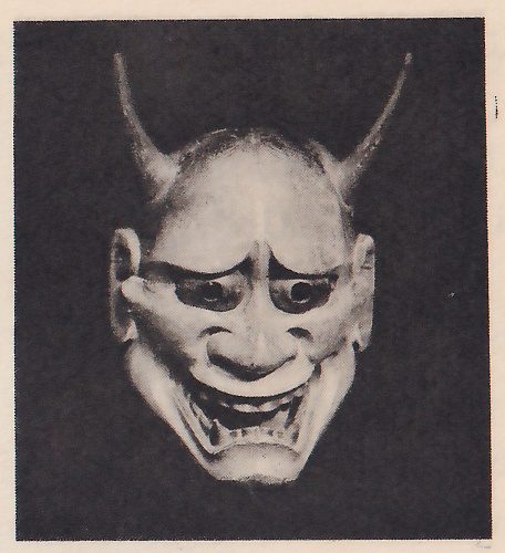 Photograph of a pale mask on a dark ground: two curved horns rising from the head, large round eyes with dark rings under drawn brows, a wide mouth open to show rows of teeth, and creases across the forehead.
