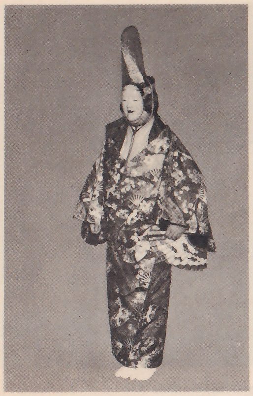 Photograph against a plain gray ground of a standing masked figure in a very tall, narrow black headdress, a wide-sleeved robe patterned with fans and flowers and white split-toe socks; a fan with a scalloped edge shows low at the viewer's right.