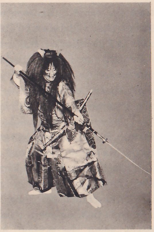 Photograph against a plain gray ground of a figure in a wide stance in a patterned robe and wide trousers patterned in light and dark, with a pale grinning mask framed by very long loose dark hair and a small ornament on top of the head; a long thin pole slants across the body from the upper left to the lower right, and a sword hangs at the waist.