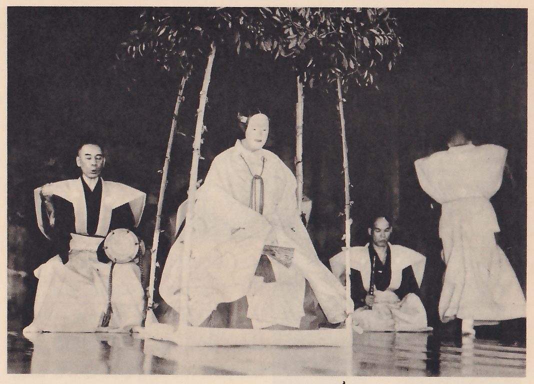 Photograph of a stage lit against a dark ground: a masked figure in a long pale robe with a cord hanging down the front sits at the center inside a frame of thin poles topped with leafy branches; at the left a seated man in a dark robe with white shoulder wings holds a small round drum at his hip, and at the right another man in the same dress sits, while a figure in a pale robe stands at the far right with the back to the viewer.
