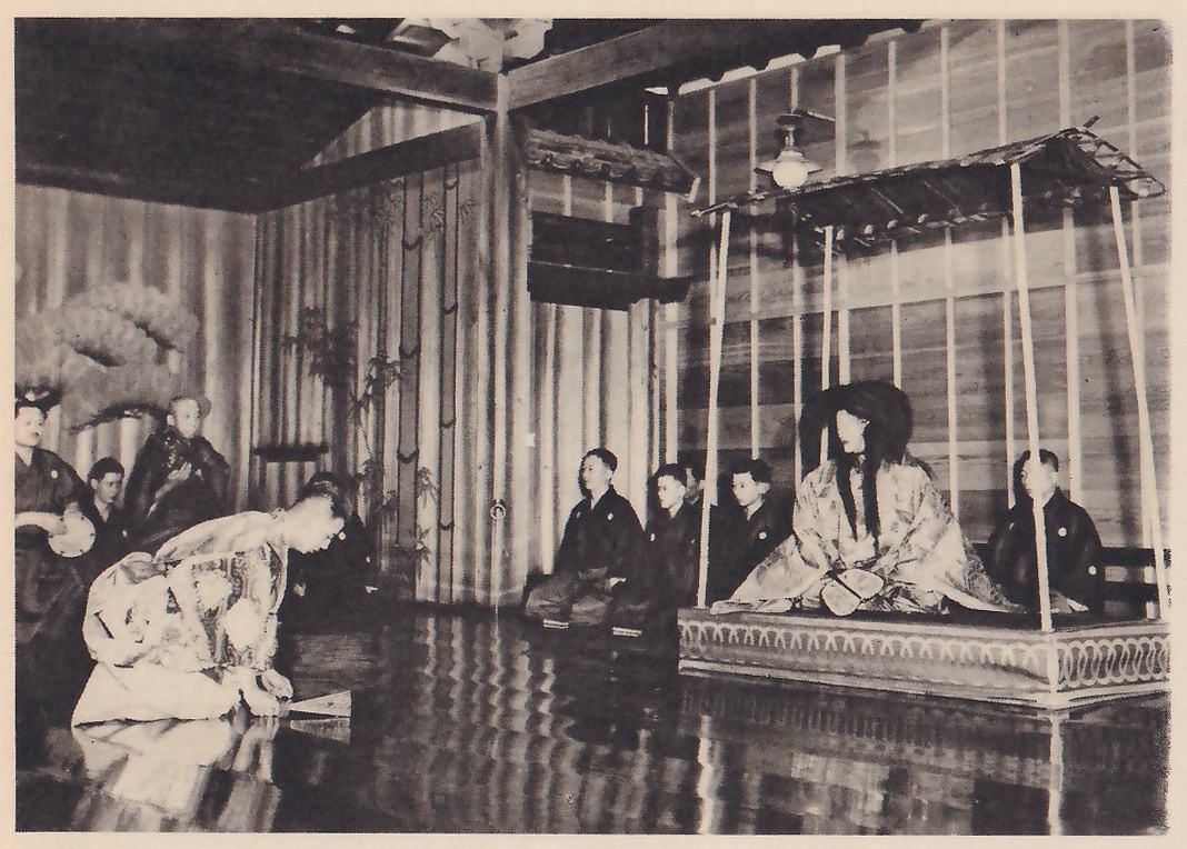 Photograph of a stage in a wooden hall: at the right, on a raised platform under a small roof with thin posts, a figure with a heavy mass of long dark hair sits in a pale robe; to the left of it several men in dark robes sit in a row on the floor, and at the far left a figure in a light patterned robe bows low with hands on the floor. A lamp hangs from the ceiling and a pine is painted on the wall at the far left.