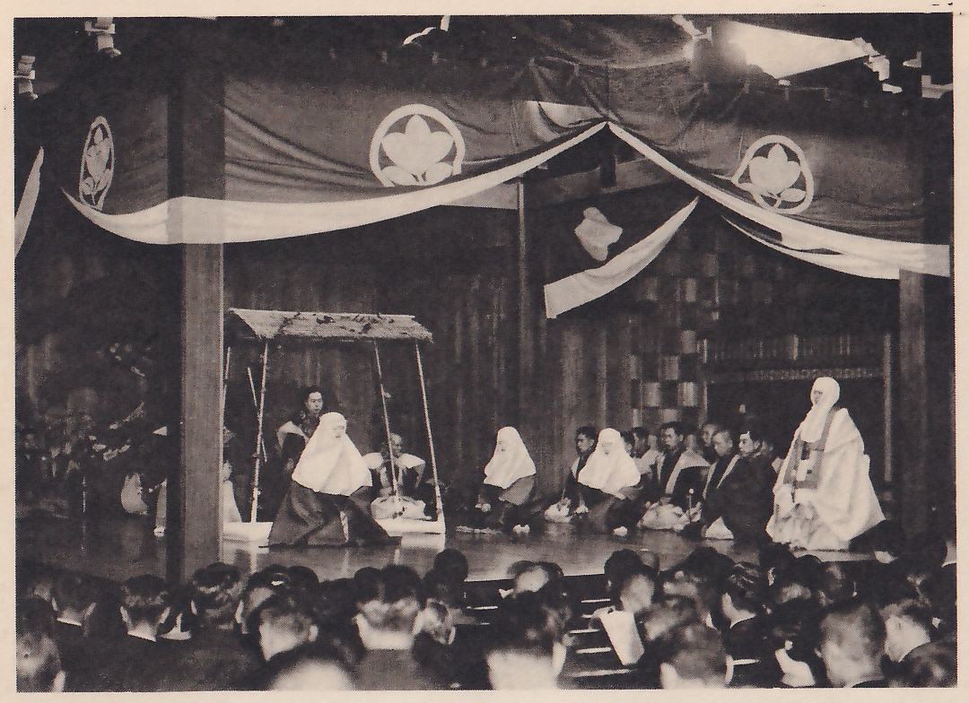 Photograph, taken from among the audience, of a stage hung with a dark cloth that carries round leaf-shaped crests, with pale bands draped across it; on the stage several figures in white head coverings and long robes sit or kneel, a small roofed frame stands at the left with a man seated inside, and a row of men in dark clothes sit at the right of center. The heads of spectators fill the bottom of the picture.