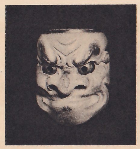 Photograph of a dark mask on a dark ground: a heavy frowning brow with curved lines above wide round eyes, a large broad nose, deep lines beside it and a wide downturned mouth above a heavy jaw.