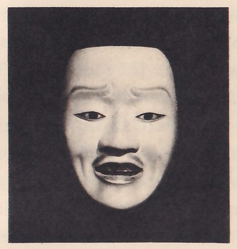 Photograph of a pale mask on a dark ground, seen from the front: a high forehead with frown creases above narrow eyes, a straight nose, a thin dark line above an open mouth, and cheeks that narrow to the chin.