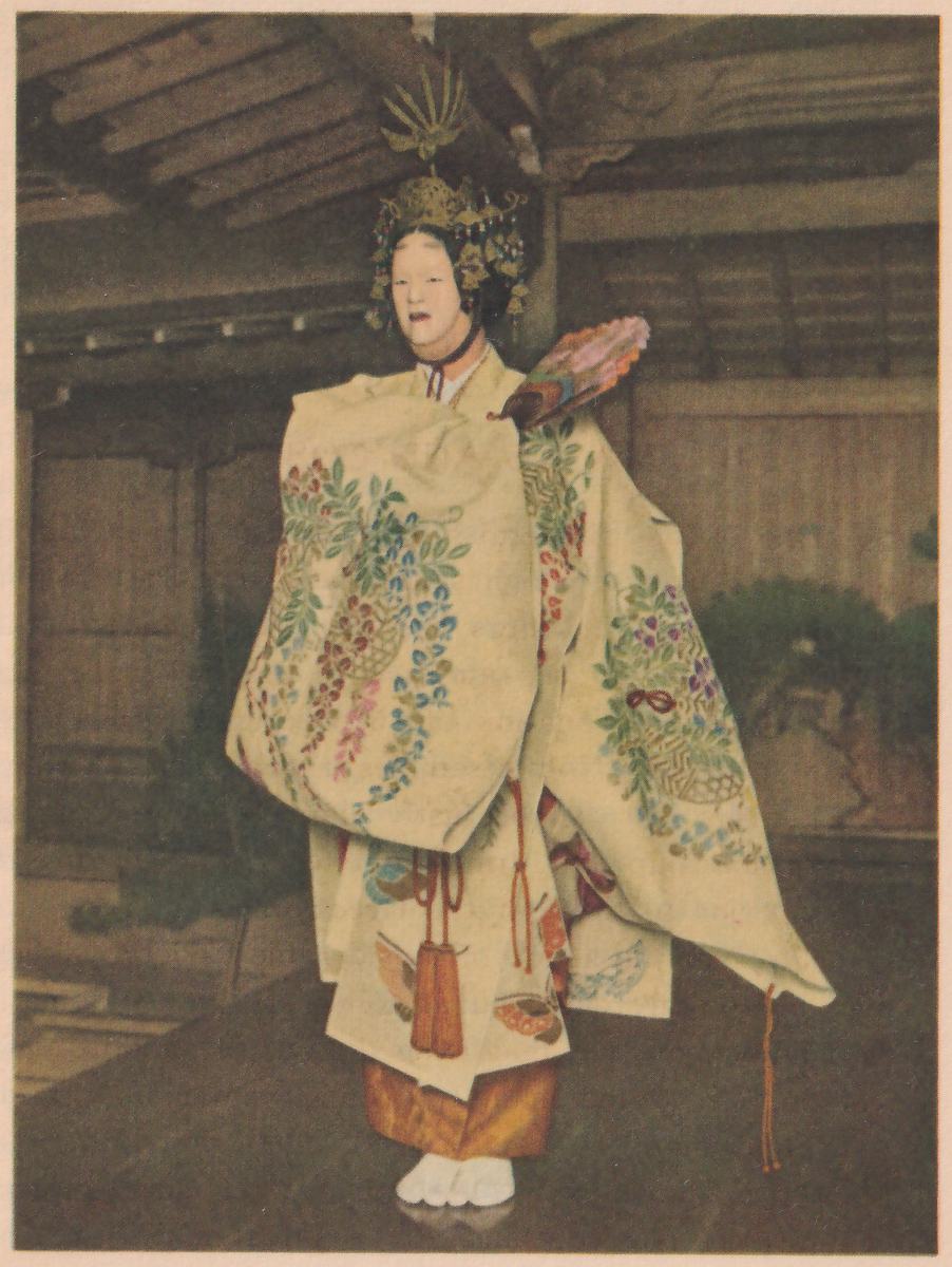 Colored photograph of a standing figure in a pale mask and a tall gold crown with hanging beads and a spiky ornament on top, in a long cream robe covered with embroidered branches and baskets in blue, green, pink and red, with a red-brown cord with tassels hanging down the front, an orange lower garment and white split-toe socks; a fan with colored decoration shows behind the shoulder at the viewer's right. The figure stands on a wooden floor in a room with a beamed ceiling and sliding panels.