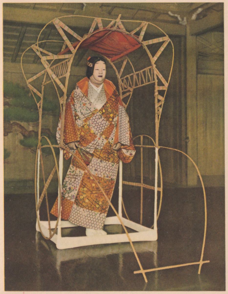 Colored photograph of a figure in a pale mask with dark hair tied with a small ornament, in an orange robe patterned with squares of white lattice and yellow and orange flowers, standing inside a large frame of thin poles with a red cloth over the top and a white base; loops of cane curve out to the right, and a fan is held low at the waist. Behind it are a dark wooden room, a pine painted on a panel at the left and a polished floor.