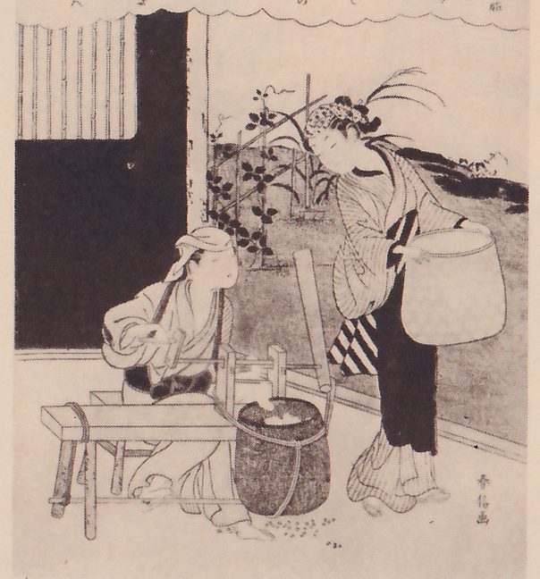 Black-and-white print of two figures outdoors beside the open front of a house. At the left a figure with a cloth tied around the head sits on a low wooden bench, working a small wooden device with a handle that stands over a rope-bound tub. At the right a second figure in a striped robe stands bending slightly forward, holding a large round basket out at arm's length. Behind them plants climb a trellis, and a dark open doorway with a blind of thin slats stands at the left. A line of small Japanese characters runs along the top edge, and a signature column stands at the lower right.