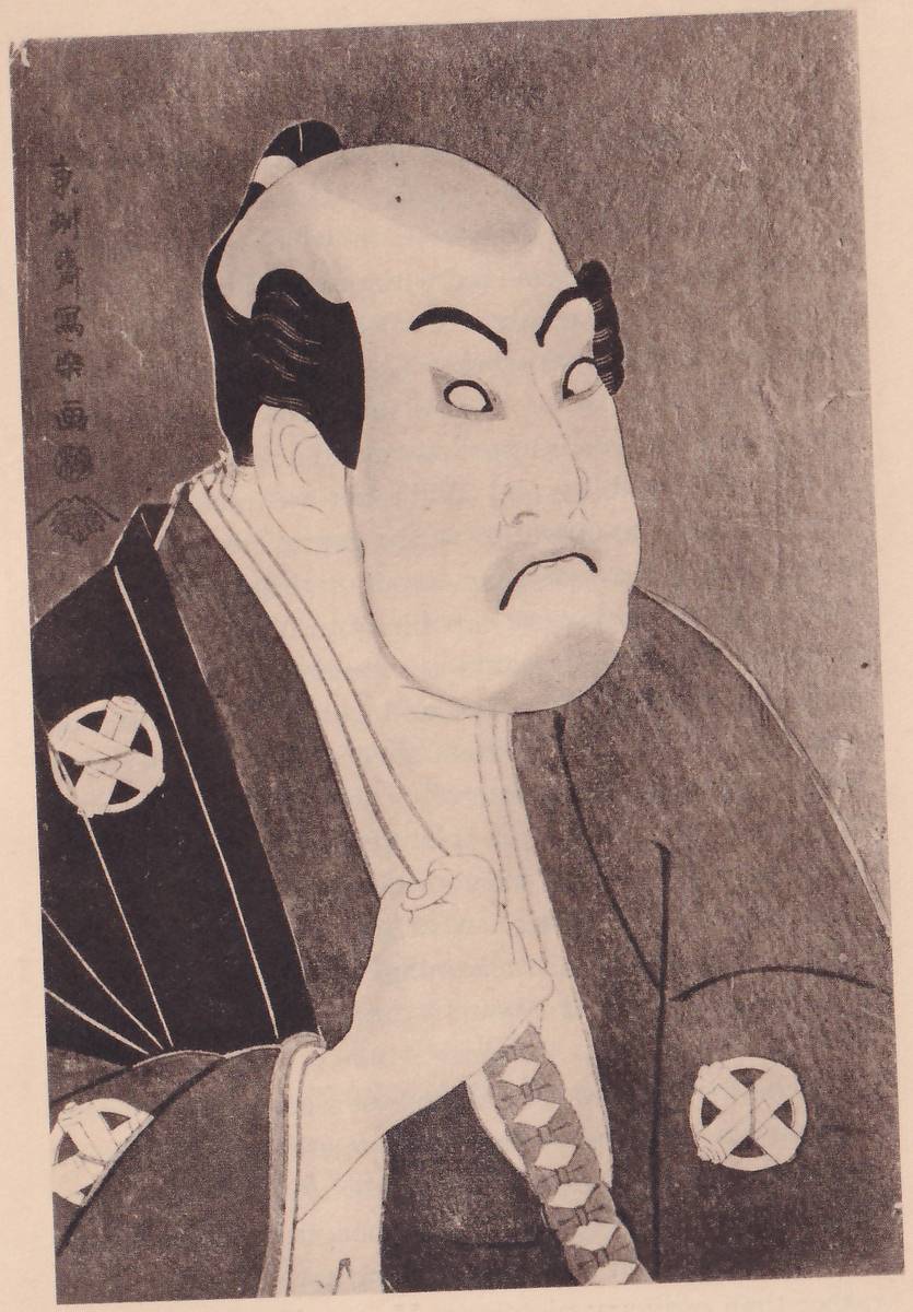 Close black-and-white print of the head and upper body of a figure with a broad face turned to the right, the brows thick and arched, the eyes turned sideways so that the whites show, and the mouth turned sharply down. The head is shaved at the top with a small knot at the back. The jacket has wide shoulders with round crests, and a fist is held in front of the chest, gripping a pale band. The background is plain and dark. A column of Japanese characters stands at the upper left, with a seal below it.