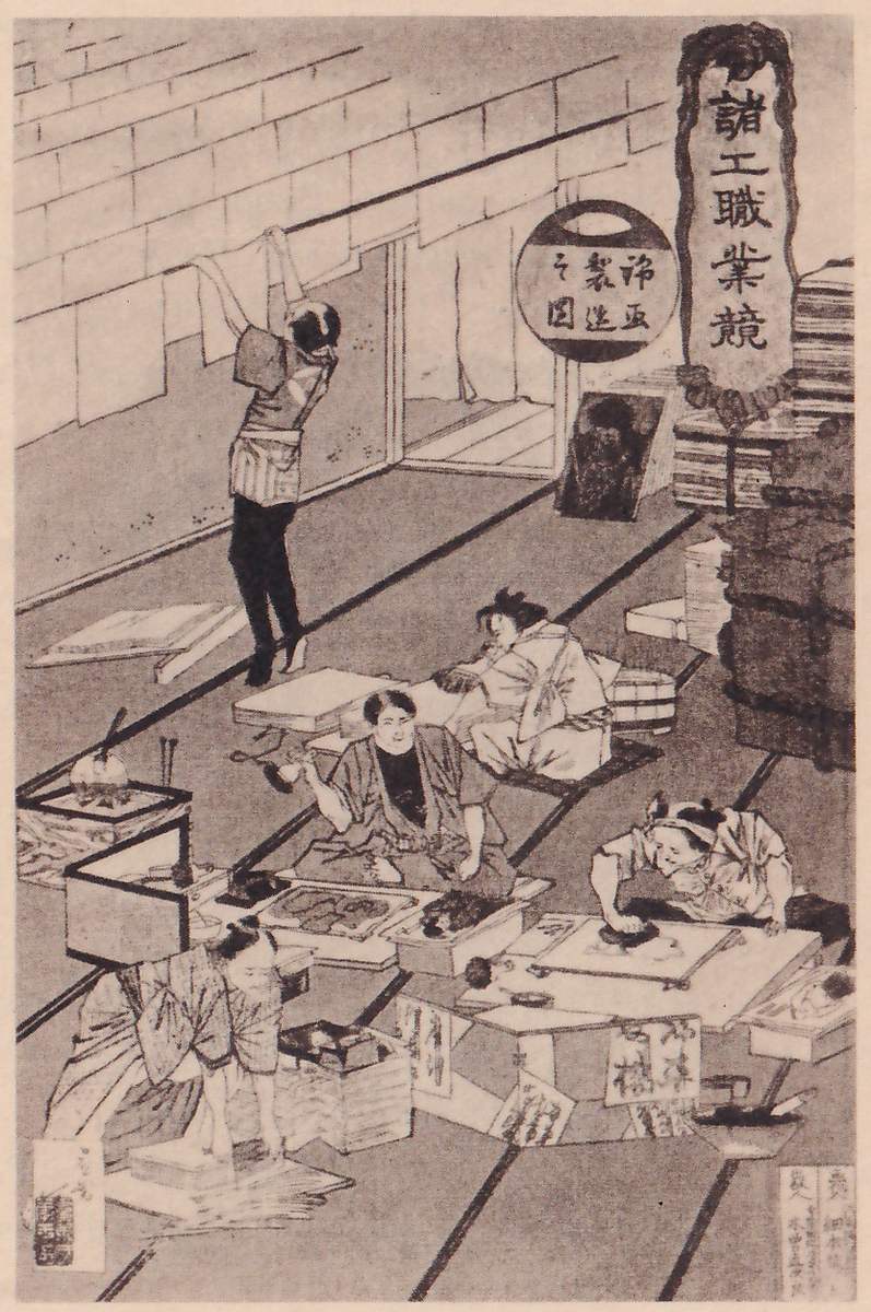Black-and-white print of a room with a floor laid in long strips and several figures at work. At the upper left a figure stands with both arms raised, hanging pale sheets along a wall of rectangular blocks. Others sit or kneel at low tables with sheets, boxes and tools, at the left, center and lower right, one bending over a table at the lower left. Stacks of flat bundles stand along the right edge. A round sign and a tall banner of Japanese characters hang at the upper right, and labels are pasted on the boxes in the foreground.