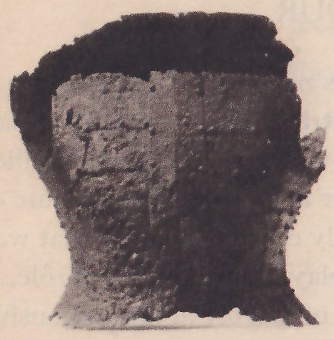 Black-and-white photograph of a rough dark object shaped like a short waisted cylinder that flares toward the top and bottom, with a jagged dark upper edge and a pitted surface. A vertical line runs down its center.