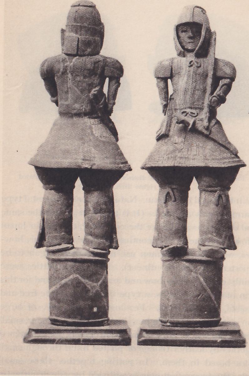 Black-and-white photograph of two upright figures side by side, each standing on a drum-shaped base set on a plinth with stepped edges. The figure at the left shows no face: its head covering has a flat panel at the back, and it has square shoulder pieces, arms bent at its sides, a flared skirt and two legs. The figure at the right has a face looking out from a hood-like head covering, square shoulder pieces, one hand on its hip, a long slanting piece beside the hip, a flared skirt and legs with small knotted loops at the knees. Faint lines cross the drum bases.