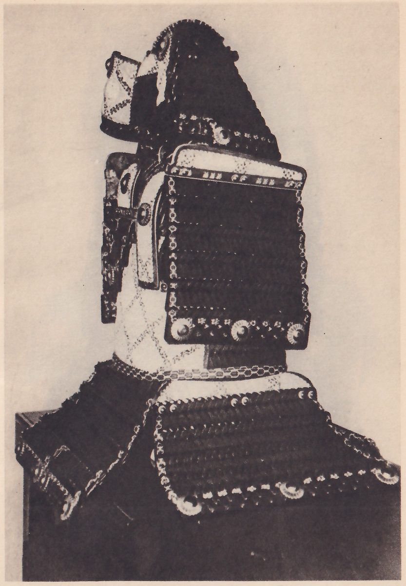 Black-and-white photograph of a suit of armour on a stand against a pale wall. A tall dark helmet with a long pointed back rises at the top, with a small pale panel patterned in diamonds hanging at its left. Below it a square panel of dark laced bands with a pale top edge and three round studs along its bottom edge stands upright, and a pale diamond-patterned section lies beneath it. At the bottom a broad dark curved piece of laced bands spreads across the width of the picture, edged with small round studs.
