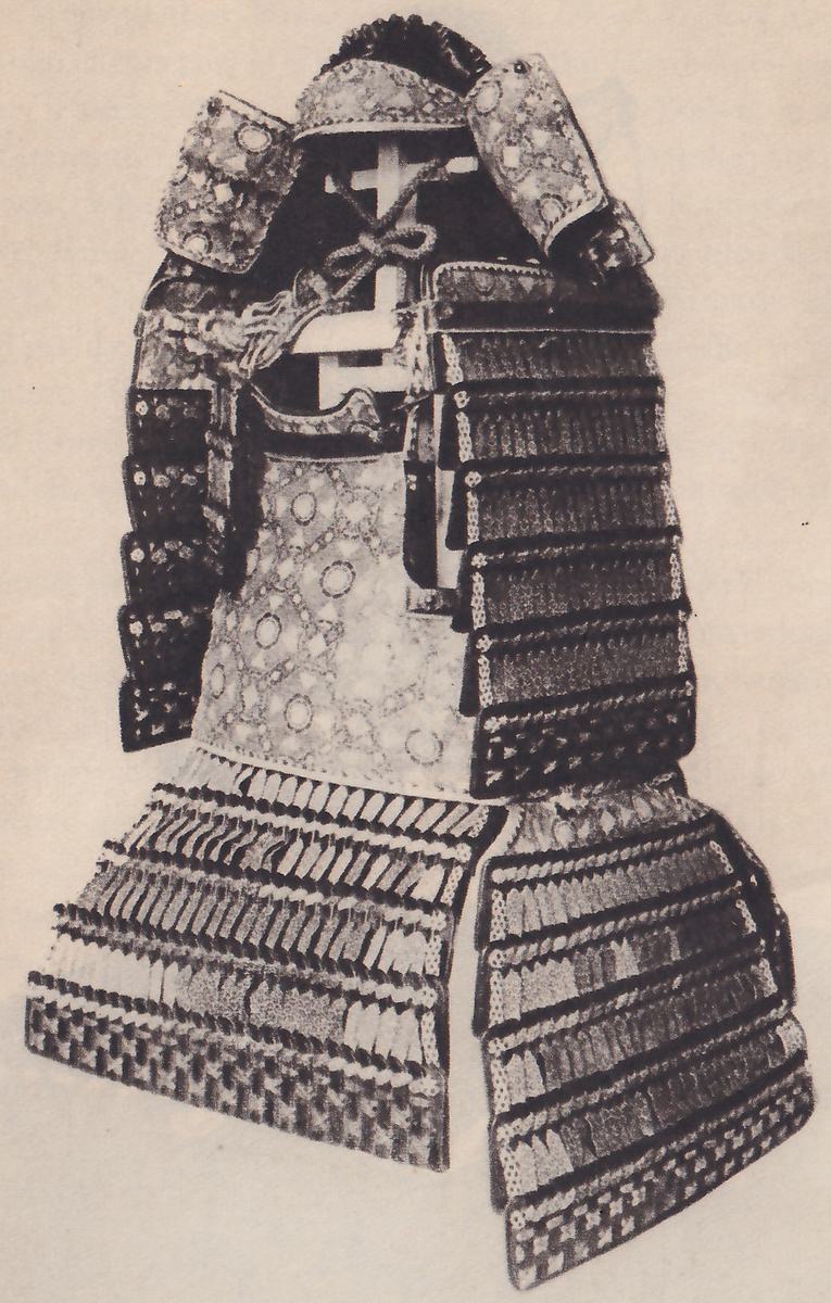 Black-and-white photograph of a suit of armour on a stand. Two broad patterned flaps stand up at the top on either side of a pale cross-shaped frame with a knotted cord. A pale body section patterned with rings and small diamonds fills the middle. Broad dark laced bands run down both sides, and two wide skirt pieces of short pale-tipped bands, each ending in a bottom row of small checks, hang at the bottom.