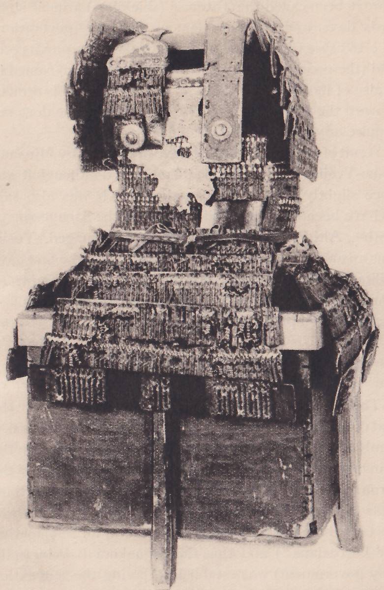 Black-and-white photograph of a badly worn, broken mass of armour set on a dark box with a flat upright post at the center of its front. Broad horizontal bands, rough and pitted, spread across the front of the box. Above them a narrower upright section has a pale ragged patch at its center and two flat rectangular plates with round discs standing at the top, with torn pieces hanging at the upper left and upper right.