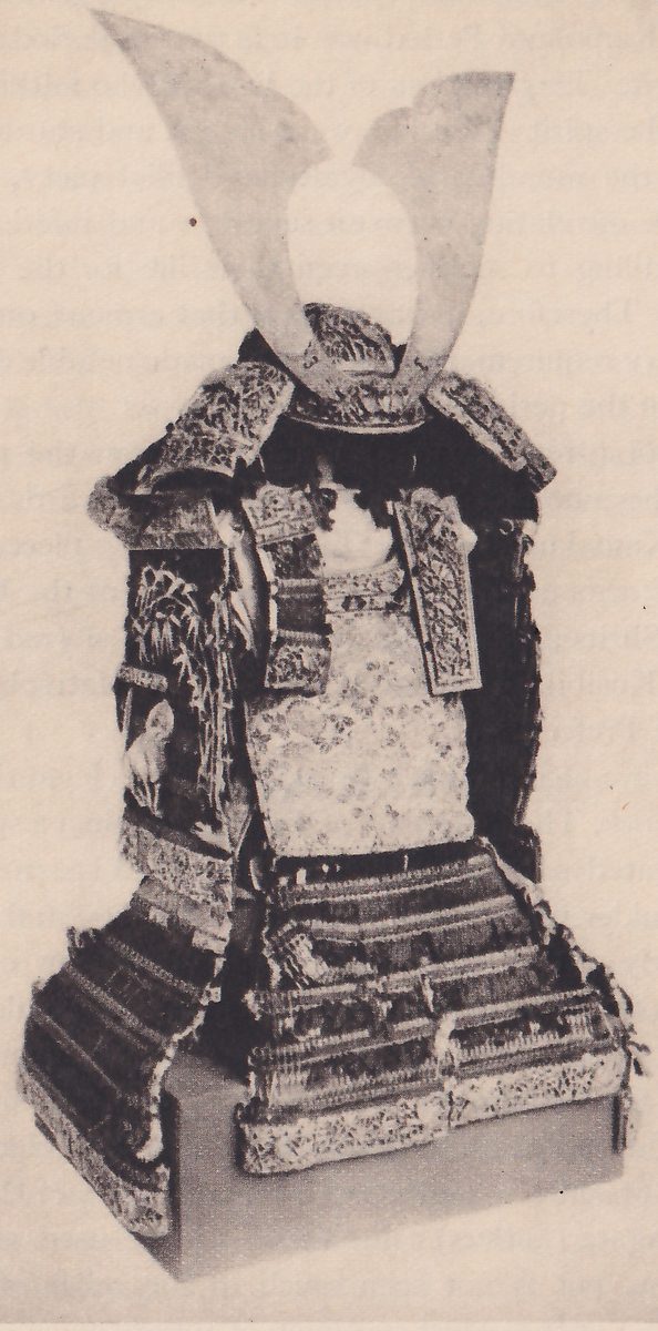 Black-and-white photograph of a suit of armour set on a low pale block. Two broad pale curved plates rise in a V from a helmet at the top. Wide shoulder pieces covered in fine patterns spread to either side below it. A dark panel at the left side carries pale pictured designs, including tall slender leaves and a rounded pale shape below them. A pale patterned front panel lies at the center, and wide skirt pieces of laced bands with patterned lower edges flare out at the bottom.