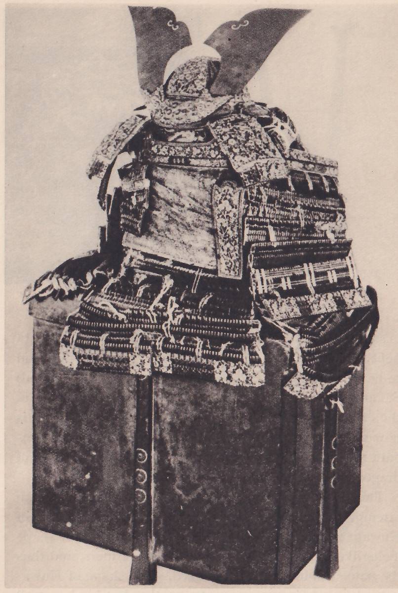 Black-and-white photograph of a suit of armour set on a tall dark box, with three small round fittings in a column on a post at the box's left. Two dark curved plates with small scroll-shaped notches rise in a V above a helmet at the top. Wide patterned shoulder pieces spread below it, a plain mottled panel lies at the center, and skirt pieces of laced bands with small pale cord ties hang over the top of the box. A heavy dark cord loops down at the right.
