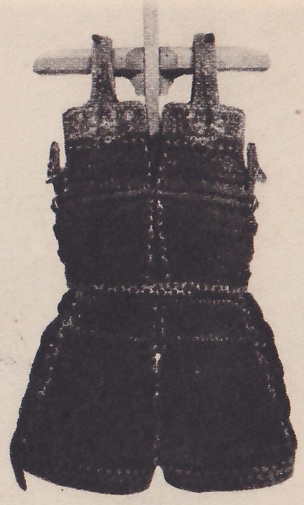 Black-and-white photograph of a dark laced body piece hanging by two upright straps from a horizontal bar on a central post. A pale patterned band lies across its top, small pale-edged stitching runs in rows across it, and a small projecting piece shows at each side of its middle. The lower part spreads out into a short skirt with a seam down the center.