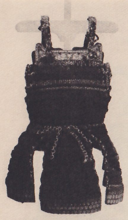 Black-and-white photograph of a dark laced body piece hanging by two narrow upright straps from a horizontal bar on a central post. A pale patterned band lies across its top. A loop of pale-edged cords crosses its lower middle, and the lower part spreads out at both sides, with a short banded piece at the lower right.